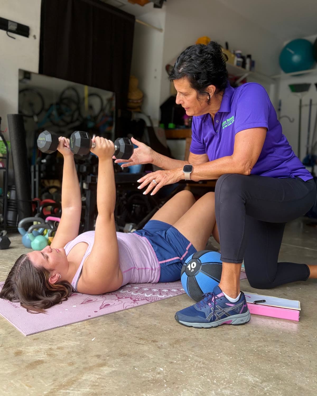 Nikki Van Culin kneeling beside a client on a mat, coaching her through a dumbbell press